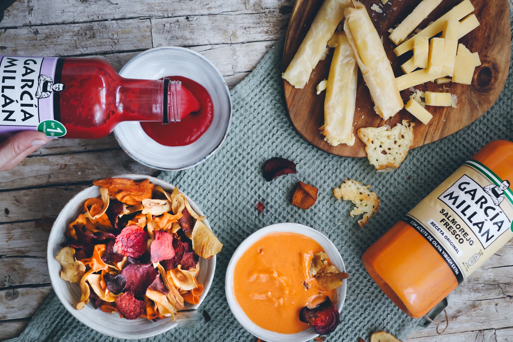 Imagen desde arriba de una mesa con chips vegetales yalgunos palillos de queso servidios con salmorejo
